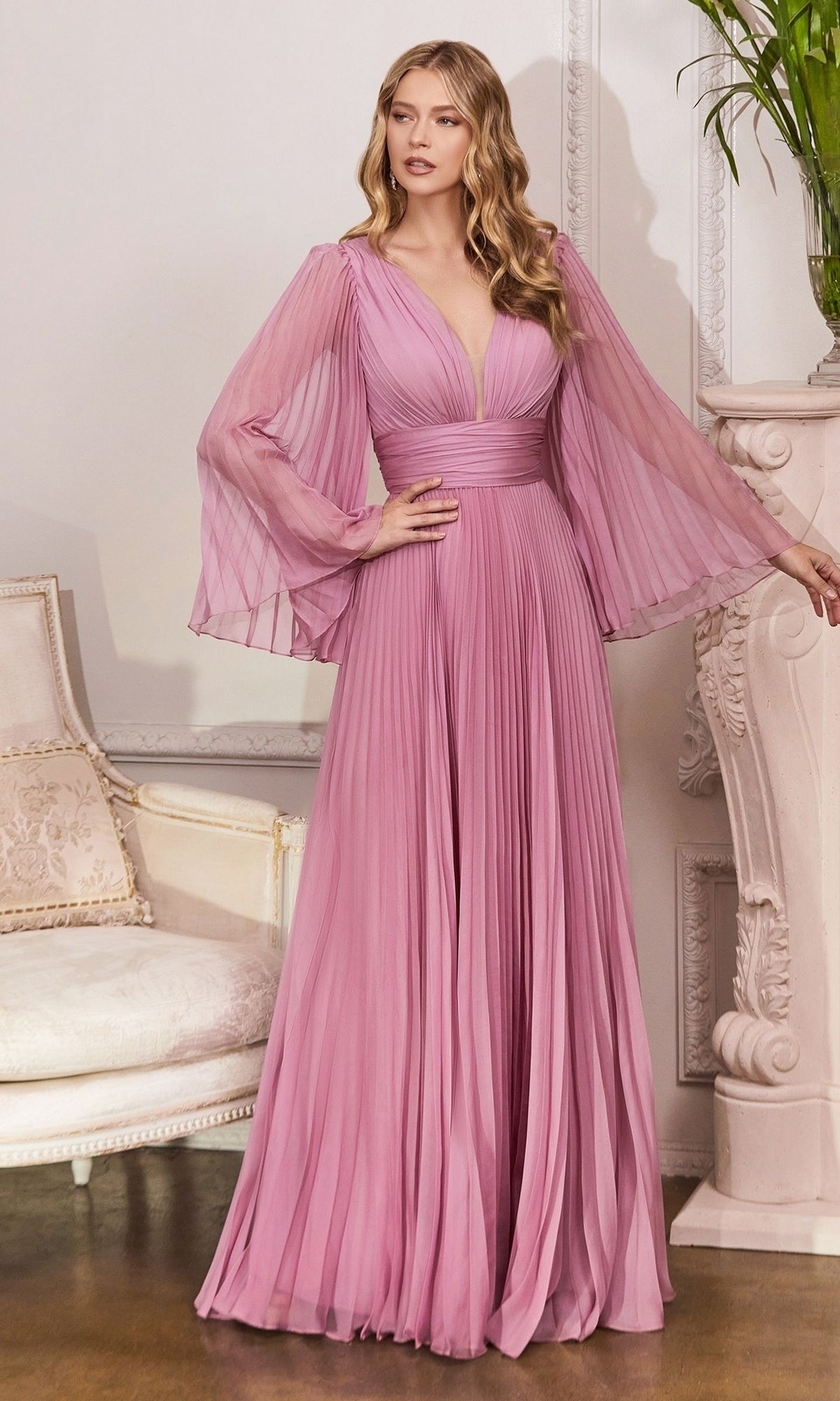 A woman stands indoors wearing the Long Bell-Sleeve Pleated Long Formal Dress CD242 in Blossom Pink, featuring sheer bell sleeves and a deep V-neckline—a perfect choice for elegant evening occasions.